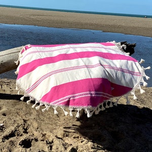 May include: A pink and white striped round beach towel with white fringe. The towel is laying on a sandy beach.