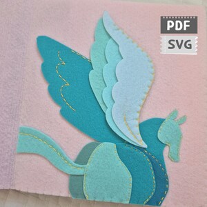 Peut inclure: Un motif Pégase en feutre dans les tons de bleu et de turquoise, avec des détails de couture jaunes. Le Pégase est posé sur un fond rose. L'image comprend le texte "PDF" et "SVG".