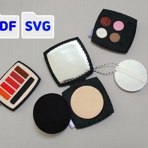 Puede incluir: Colección de juguetes de maquillaje de fieltro, que incluye un espejo compacto, un compacto de polvos, una paleta de sombras de ojos y una borla. La paleta de sombras de ojos presenta tonos rojos, naranjas y marrones. La imagen también incluye el texto "PDF SVG".