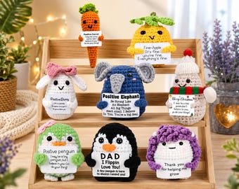 May include: A selection of handmade crochet figures, including a carrot, pineapple, bunny, elephant, snowman, penguin, and sheep. Each figure holds a sign with an inspirational message. The figures are displayed on a wooden shelf.