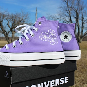May include: Purple high-top Converse sneakers with white floral designs and a white platform sole. The shoes are on a black box with the Converse logo.