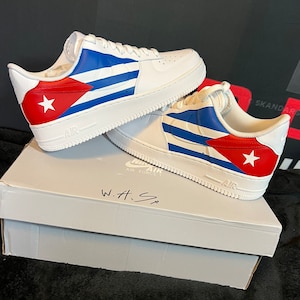May include: White leather sneakers with a Cuban flag design. The sneakers have blue and red stripes with a white star on the red stripe. The sneakers are sitting on a white box with the words "Air Force 1" and "W.H.S" written on it.
