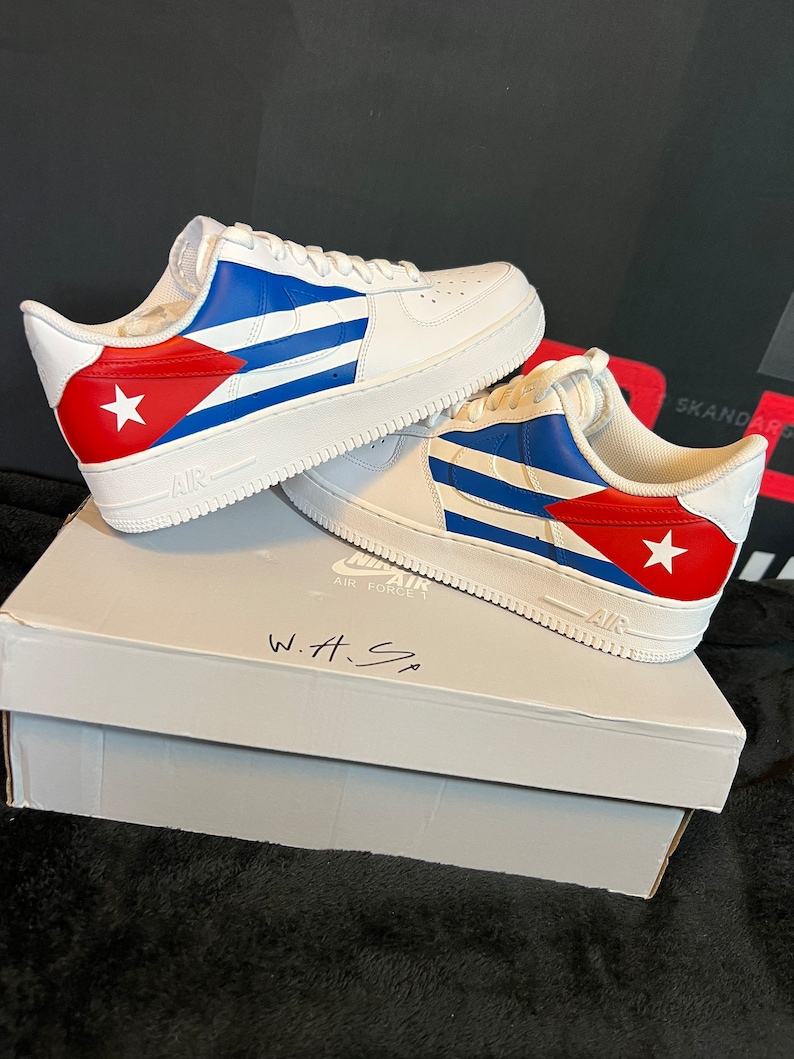 May include: White leather sneakers with a Cuban flag design. The sneakers have blue and red stripes with a white star on the red stripe. The sneakers are sitting on a white box with the words "Air Force 1" and "W.H.S" written on it.