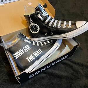 May include: A pair of black leather Converse high-top sneakers with white laces and a white rubber sole. The sneakers have the text "Sorry for the wait" printed on the side in white lettering. The sneakers are in a box with the Converse logo.