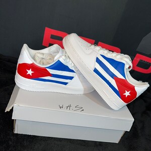 May include: White sneakers with a Cuban flag design. The sneakers are sitting on a white box with the words "W.H.S" written on it.