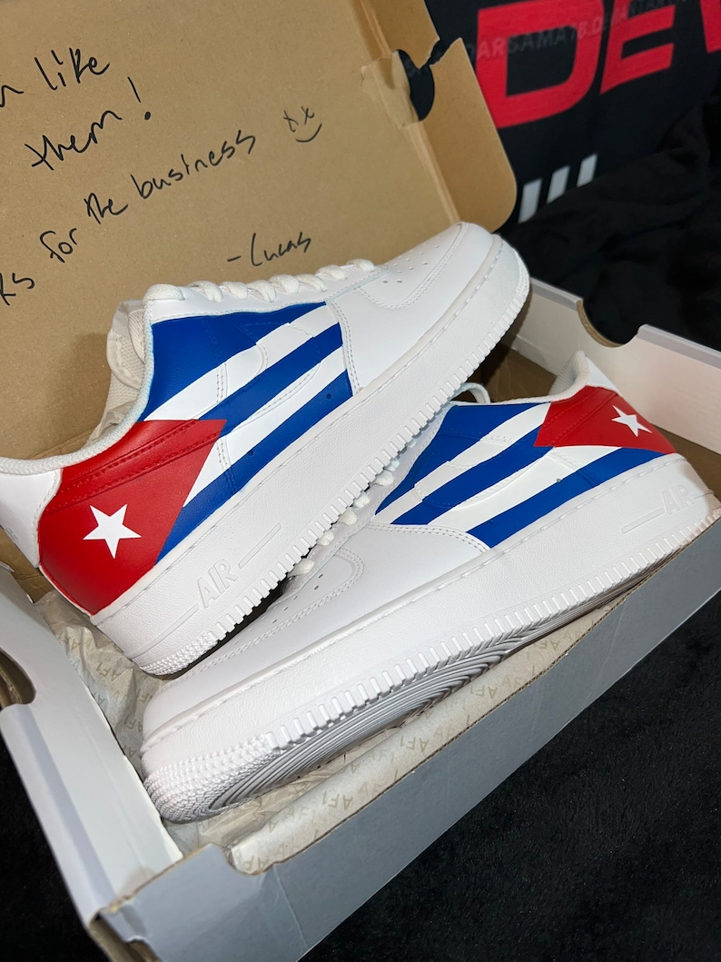 May include: A pair of white sneakers with a custom design featuring the Cuban flag colors: red, white, and blue. The sneakers are in a shoebox with a handwritten note that says "I like them! Thanks for the business - Chels"