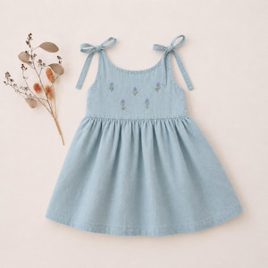 May include: Light blue denim dress with tie straps and a gathered skirt. The bodice features embroidered purple floral accents. A sprig of dried flowers rests to the left of the dress.