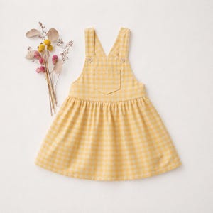 Puede incluir: Un vestido peto de cuadros amarillos y blancos con tirantes ajustables y un bolsillo delantero. El vestido tiene una falda fruncida y está adornado con dos pequeños botones en los tirantes. Un pequeño ramo de flores secas se coloca a la izquierda del vestido.