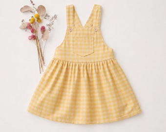 May include: A yellow and white gingham-patterned pinafore dress with adjustable straps and a front pocket. The dress has a gathered skirt and is adorned with two small buttons on the straps. A small bouquet of dried flowers is placed to the left of the dress.