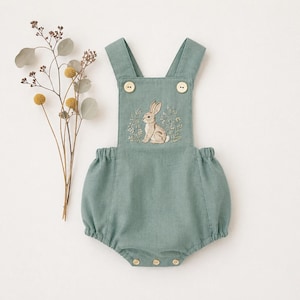 May include: A sage green baby romper with an embroidered bunny design. The romper has adjustable straps with wooden buttons and a gathered waist. The bunny embroidery is surrounded by delicate floral details. The garment is made of soft fabric.