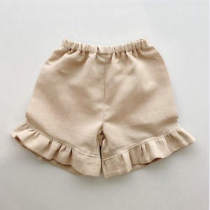 May include: A pair of tan shorts with an elasticated waistband and a frilled hem. The shorts are made from a light fabric. These shorts are suitable for warm weather and casual wear. The shorts are a neutral colour.