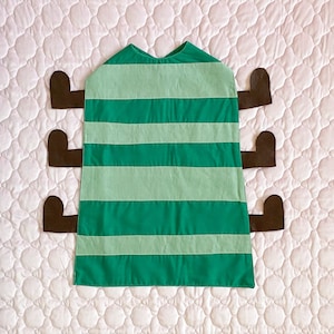 Peut inclure: Une cape en tissu rayée verte et vert clair avec des bras en feutre marron attachés sur les côtés. La cape est conçue pour ressembler à une chenille.