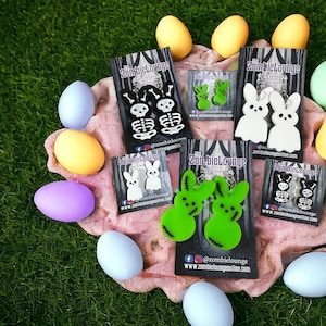 Puede incluir: Surtido de pendientes en forma de huevo de Pascua de colores con varios diseños, incluyendo esqueletos, conejos y peeps verdes. Los pendientes se muestran en una tela rosa y blanca con el texto "ZombieLounge" y sus identificadores de redes sociales.