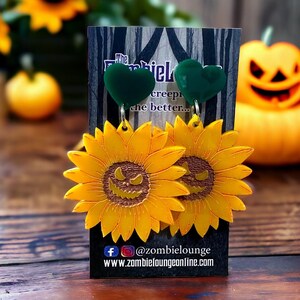 May include: A pair of yellow sunflower earrings with a spooky jack-o'-lantern face in the centre. The earrings are attached to green heart-shaped charms. The earrings are displayed on a black card with the text "The creepier the better..." and a social media handle.
