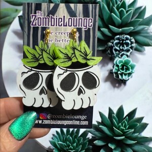 May include: A pair of white skull earrings with green plant accents. The earrings are on a card with the text "The Zombielounge" and "@zombielounge" and "www.zombieloungeonline.com".