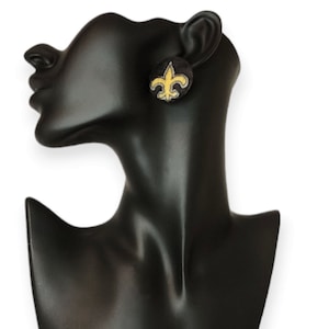May include: A pair of black and gold stud earrings featuring the fleur-de-lis logo of the New Orleans Saints.