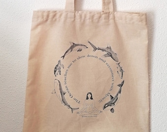 May include: A beige tote bag with a black and white illustration of a person meditating surrounded by ocean animals. The text reads "The ocean teaches me to slow down and reconnect with myself".