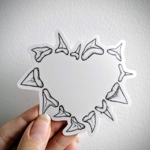 May include: A white sticker in the shape of a heart, outlined with black shark teeth. The teeth are detailed with black stippling. The sticker is held against a white background.