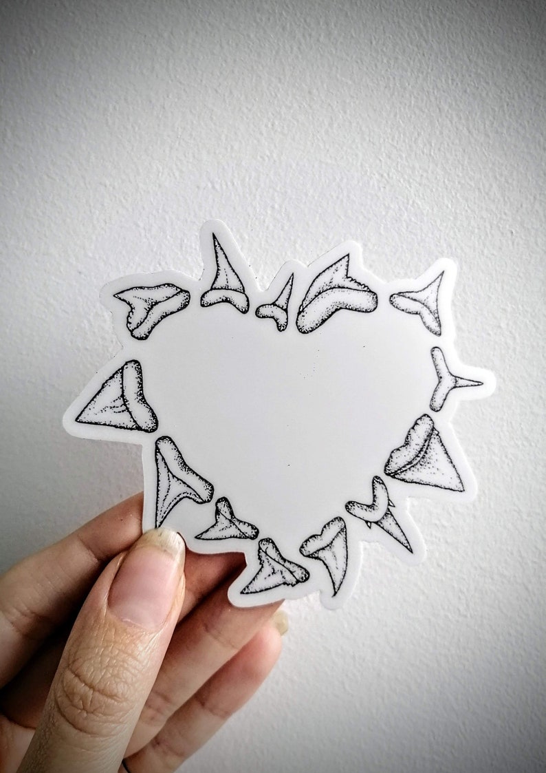 May include: A white sticker in the shape of a heart, outlined with black shark teeth. The teeth are detailed with black stippling. The sticker is held against a white background.