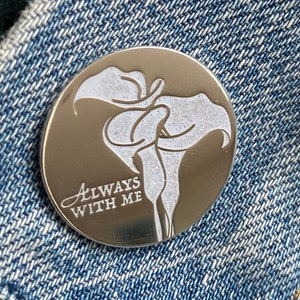 Puede incluir: Un pin de plata con un diseño de flor blanca y el texto "ALWAYS WITH ME".
