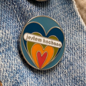 May include: A blue enamel pin featuring a multi-coloured heart design. The heart has a red centre and is surrounded by orange, yellow and green layers. The pin has the text "jestem kochana" in white letters on a green background.