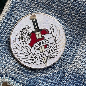 May include: A silver pin with a white background and glitter. The pin features a red heart with a silver dagger through it. The dagger has a black handle and a silver blade. The heart has a banner that reads "Always With Me". The heart is surrounded by silver wings.