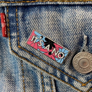 Op de afbeelding: Een blauwe, roze en witte emaille pin met een zwarte bloem en de tekst "It's a no" op een spijkerjack.