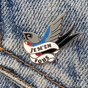 Puede incluir: Un pin de esmalte plateado con un pájaro estilizado negro y gris con detalles azules. El pájaro sostiene una pancarta blanca con el texto "JE M'EN FOU" en negro y rojo.