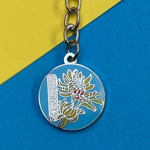 May include: A silver keychain with a blue circular charm featuring a yellow and red flower with the Japanese text "いってらっしゃい" on a white rectangle.