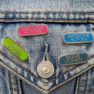 May include: Four enamel pins on a blue denim jacket. The pins are rectangular and have silver metal frames. The pins read "Fabulous Human", "Super Human", "Awesome Human", and "Brave Human". The pins are in pink, green, blue, and teal.