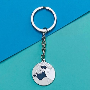 Peut inclure: Porte-clés argenté avec un charme rond représentant une vague en noir et blanc avec du texte japonais qui se lit "七転び八起き".