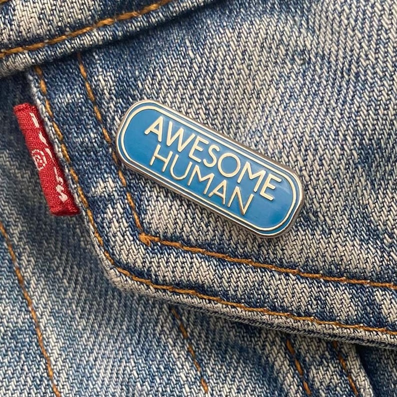 May include: A blue enamel pin with a silver border. The pin says "Awesome Human" in white letters.