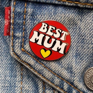 Op de afbeelding: Rode en witte emaille pin met de tekst "BEST MUM" in een retro lettertype. Een geel hart bevindt zich in het midden van de pin.