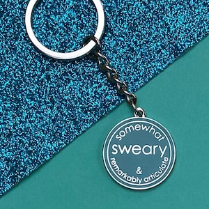 Puede incluir: Un llavero de plata con un dije redondo de esmalte azul que dice "somewhat sweaky & remarkably articulate".
