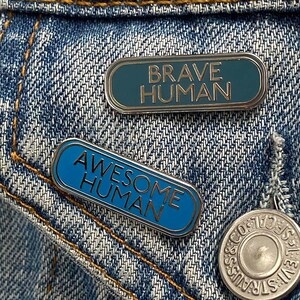 May include: Two enamel pins on a blue denim background. The pins are both oval shaped and have a silver border. The top pin says "Brave Human" in white text on a teal background. The bottom pin says "Awesome Human" in white text on a blue background.