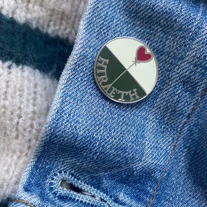 May include: A circular enamel pin with a silver edge. The pin features a design split into four sections: white, dark green, and a red heart with an arrow. The words "HIR" and "AFTH" are written in green around the edge. The pin is on a blue denim jacket.