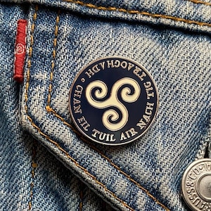 Puede incluir: Un pin redondo de esmalte azul marino con un diseño de Triskele celta blanco. El pin está inscrito con texto en un patrón circular. El pin está sujeto a una chaqueta vaquera.