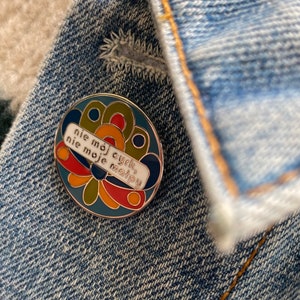 Peut inclure: Une épingle en émail colorée avec un fond blanc et le texte "nie mój cyrk, nie moje małpy" en lettres noires. L'épingle présente un motif de fleur stylisé avec des couleurs rouge, jaune, verte et bleue.