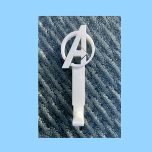 Pode incluir: Gancho de parede de pl&aacute;stico branco em forma de logotipo dos Vingadores, com um c&iacute;rculo contendo a letra "A". O gancho est&aacute; montado em uma superf&iacute;cie azul texturizada.