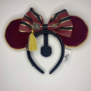 Może przedstawiać: Czerwone aksamitne uszy Myszki Minnie z złotą obwódką i czarną kokardką w złote paski. Z kokardki zwisa złoty brelok w kształcie klucza z frędzlem. Opaska na głowę jest czarna.