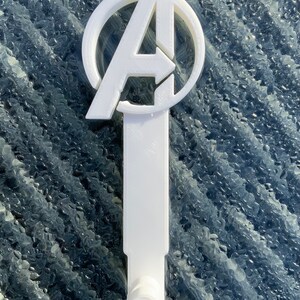 Pode incluir: Gancho de parede de pl&aacute;stico branco em forma de logotipo dos Vingadores. O logotipo &eacute; um c&iacute;rculo com um "A" estilizado dentro. O gancho &eacute; preso a uma base retangular.