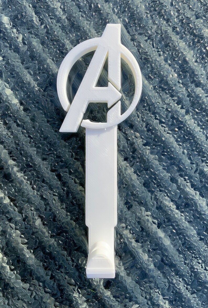 Pode incluir: Gancho de parede de pl&aacute;stico branco em forma de logotipo dos Vingadores. O logotipo &eacute; um c&iacute;rculo com um "A" estilizado dentro. O gancho &eacute; preso a uma base retangular.