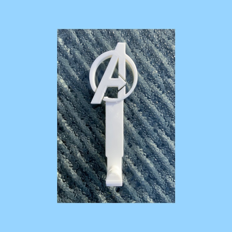 Pode incluir: Gancho de parede de pl&aacute;stico branco em forma de logotipo dos Vingadores, com um c&iacute;rculo contendo a letra "A". O gancho est&aacute; montado em uma superf&iacute;cie azul texturizada.