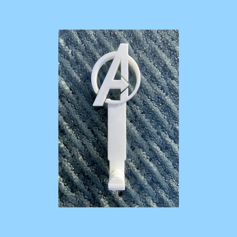 Pode incluir: Gancho de parede de pl&aacute;stico branco em forma de logotipo dos Vingadores, com um c&iacute;rculo contendo a letra "A".