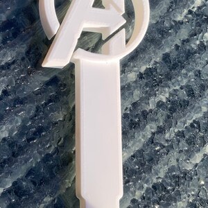 Pode incluir: Gancho de pl&aacute;stico branco com um logotipo dos Vingadores impresso em 3D. O logotipo &eacute; um c&iacute;rculo com a letra "A" dentro e uma seta apontando para a direita.