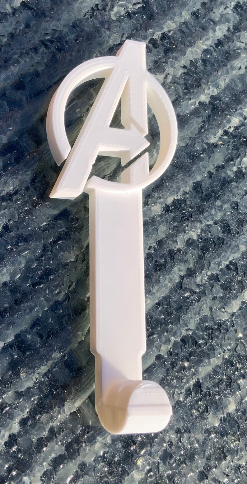 Pode incluir: Gancho de pl&aacute;stico branco com um logotipo dos Vingadores impresso em 3D. O logotipo &eacute; um c&iacute;rculo com a letra "A" dentro e uma seta apontando para a direita.