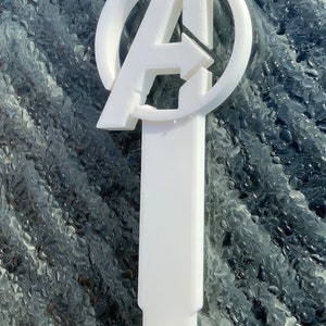 Pode incluir: Gancho de pl&aacute;stico branco em forma do logotipo dos Vingadores. O gancho &eacute; projetado para pendurar itens em uma parede ou porta.