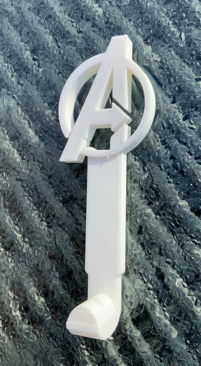 Pode incluir: Gancho de pl&aacute;stico branco em forma do logotipo dos Vingadores. O gancho &eacute; projetado para pendurar itens em uma parede ou porta.