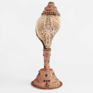 Puede incluir: Una concha dorada con intrincados tallados y piedras preciosas de colores. La concha está montada en un soporte decorativo con una base.