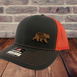 May include: A black and orange trucker hat with a brown leather patch that says "Papa Bear" and a brown bear silhouette.